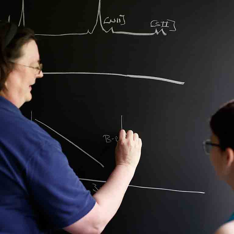 A professor writes on a chalkboard while a student watches.