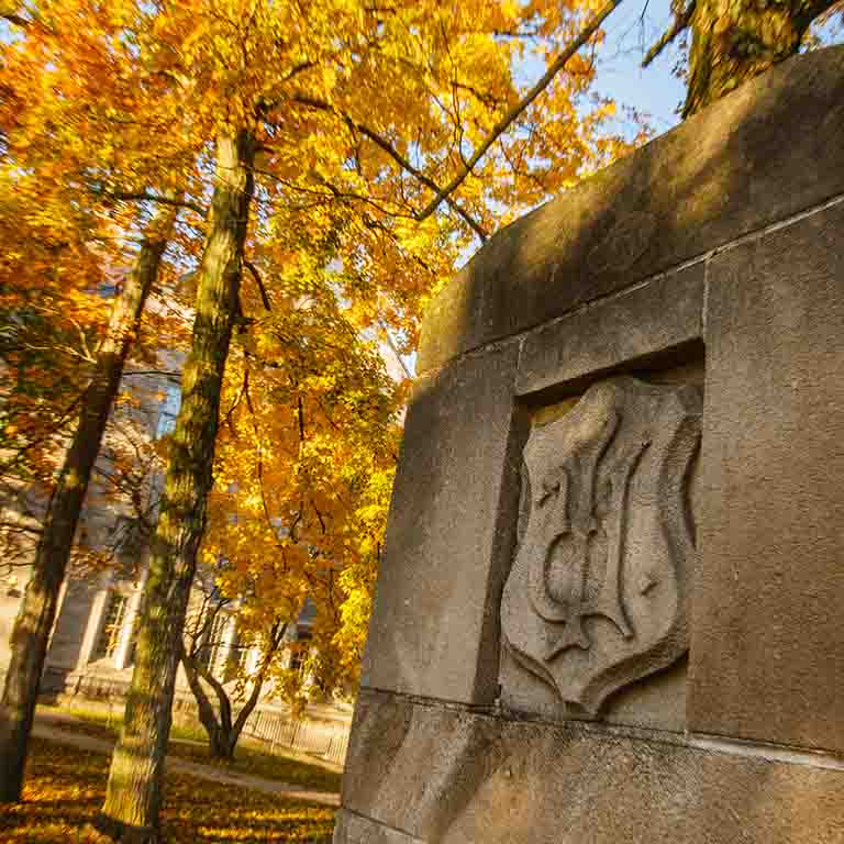 An IU emblem on a stone structure, beside trees with orange and yellow leaves in autumn