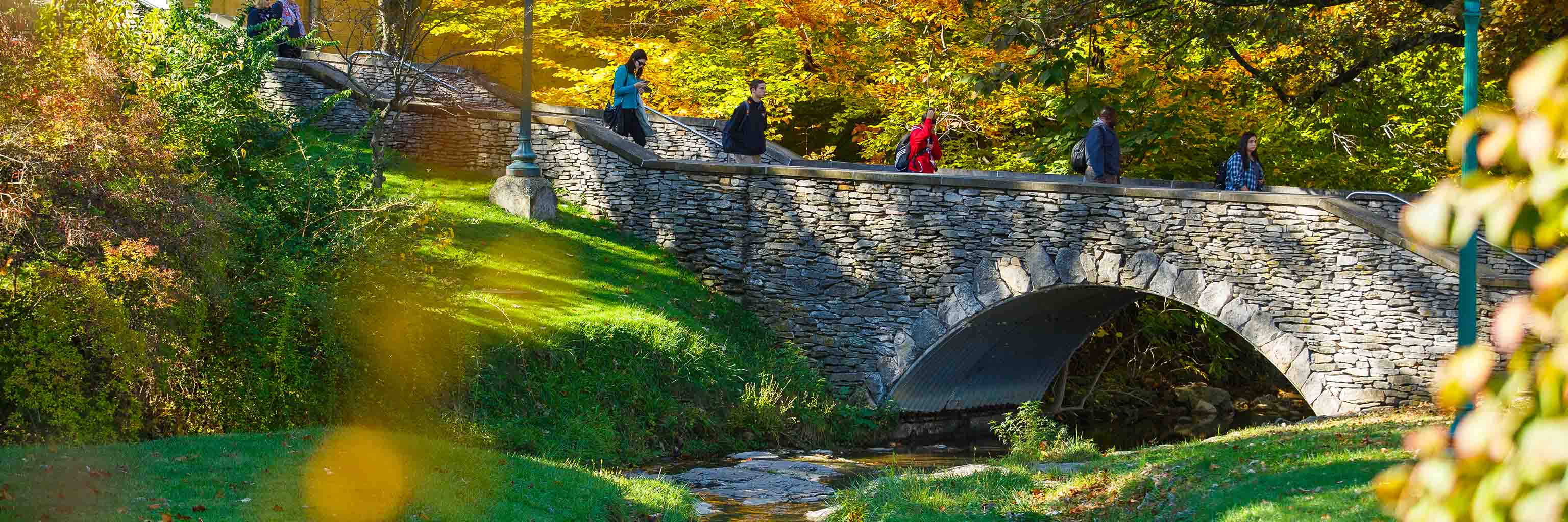 People walk across a stone bridge that spans the Campus River.