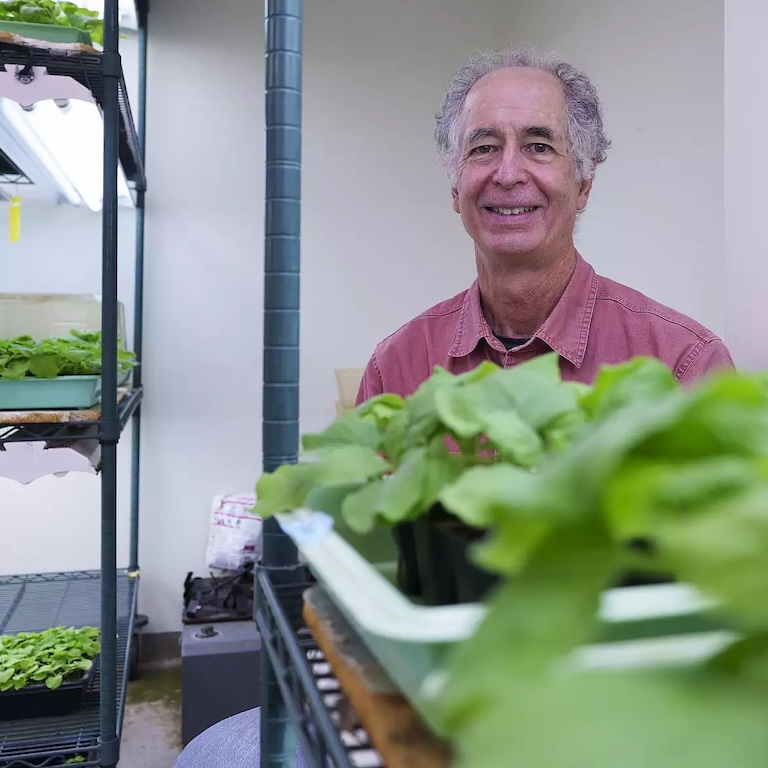 A photo of Roger Innes, who poses near various plants in a greenhouse.